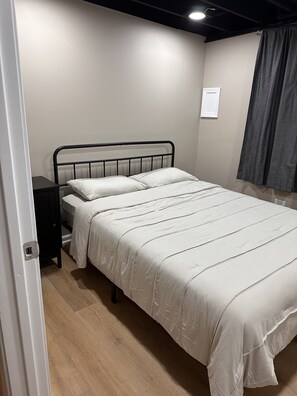 A bedroom with a single bed, a nightstand, a wall-mounted light, and a window with curtains.
