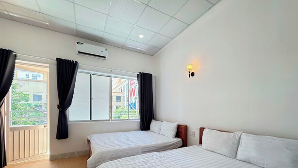 A hotel room with two beds, a window with curtains, an air conditioning unit, and a wall-mounted light fixture. - New Hotel Vung Tau (Ho Chi Minh City)