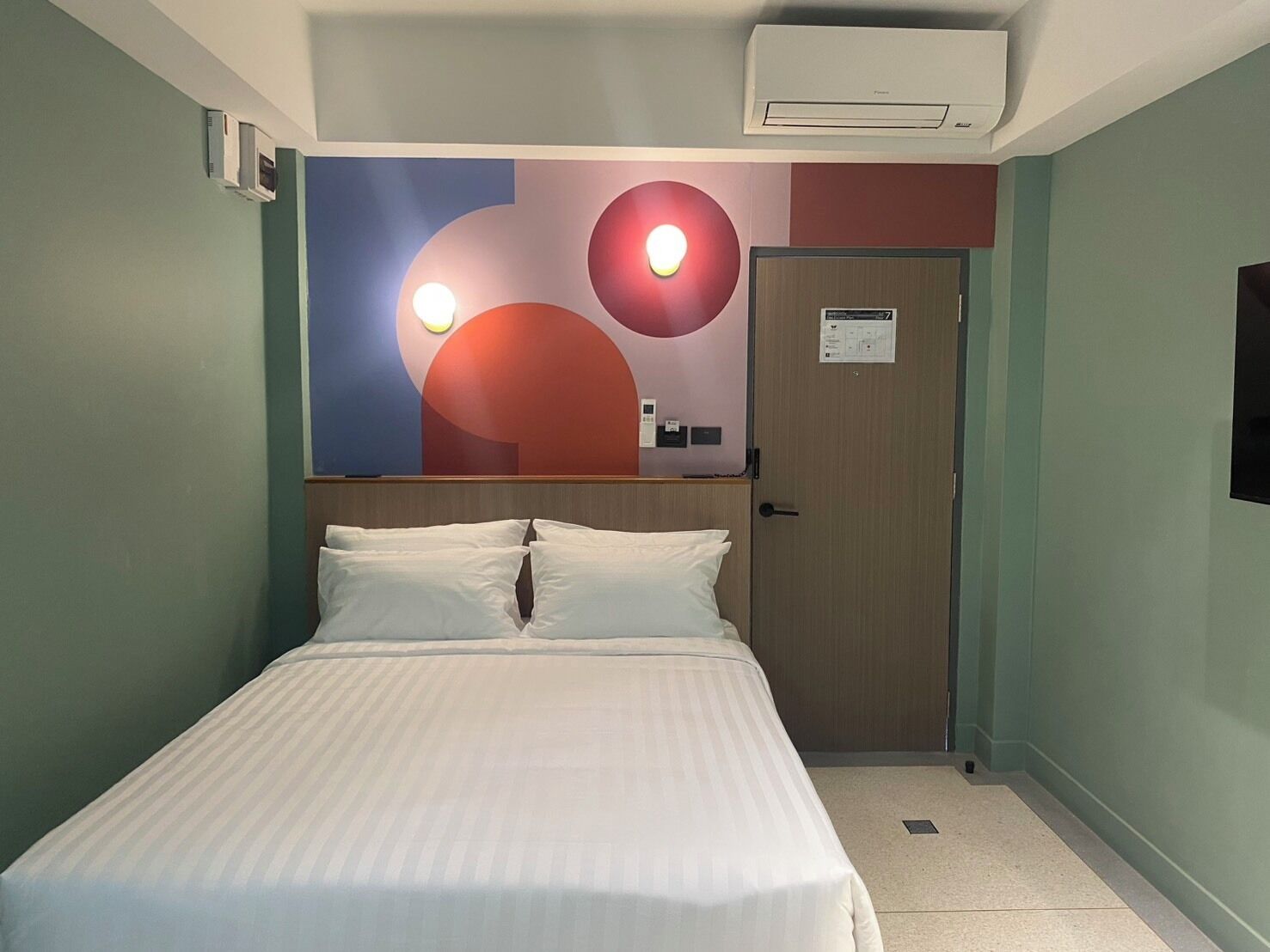 A hotel room with a bed, a wall-mounted air conditioner, and a door with a sign.