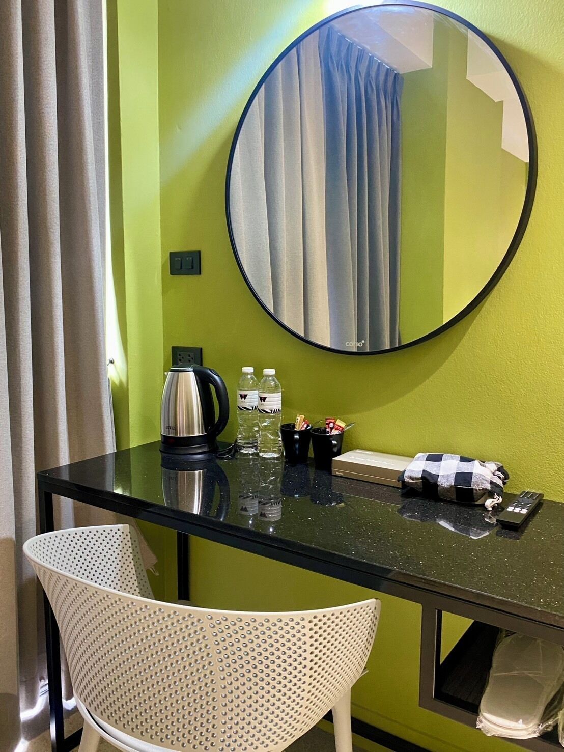 Een hotelkamer met een ronde spiegel, een tafel met waterkoker, flessen en een afstandsbediening.