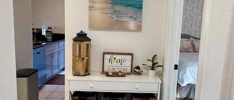 Um corredor com uma cozinha ao fundo, um quadro na parede, uma pequena mesa com uma placa e cestos de vime em uma prateleira.