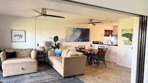 A living room with a sofa, a coffee table, and a dining area with chairs and a table. There is a ceiling fan, a plant, and a wall with a picture.