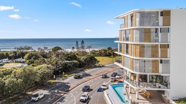 Un hôtel à plusieurs étages doté d’une piscine, offrant une vue sur la plage et donnant sur une rue bordée de voitures garées.