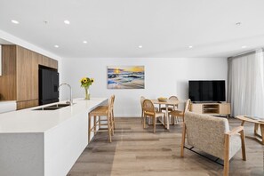 A modern kitchen with a central island, dining area with wooden chairs, a flat-screen TV, and a painting of a sunset. - Sakara @Maroochydore (Maroochydore)
