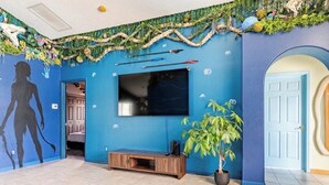 Quarto com uma televisão de tela plana grande fixada em uma parede azul, um móvel de entretenimento de madeira, uma planta em vaso e uma porta com espelho circular.