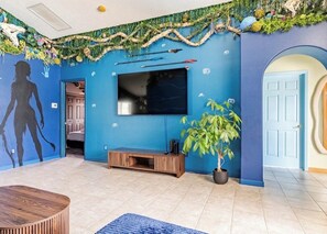 A room with a large flat-screen TV mounted on a blue wall, a wooden entertainment unit, a potted plant, and a door with a circular mirror. - Once Upon A Vacation Home with Pool + Game Room + Sleeps 9 near DISNEY! (Kissimmee)