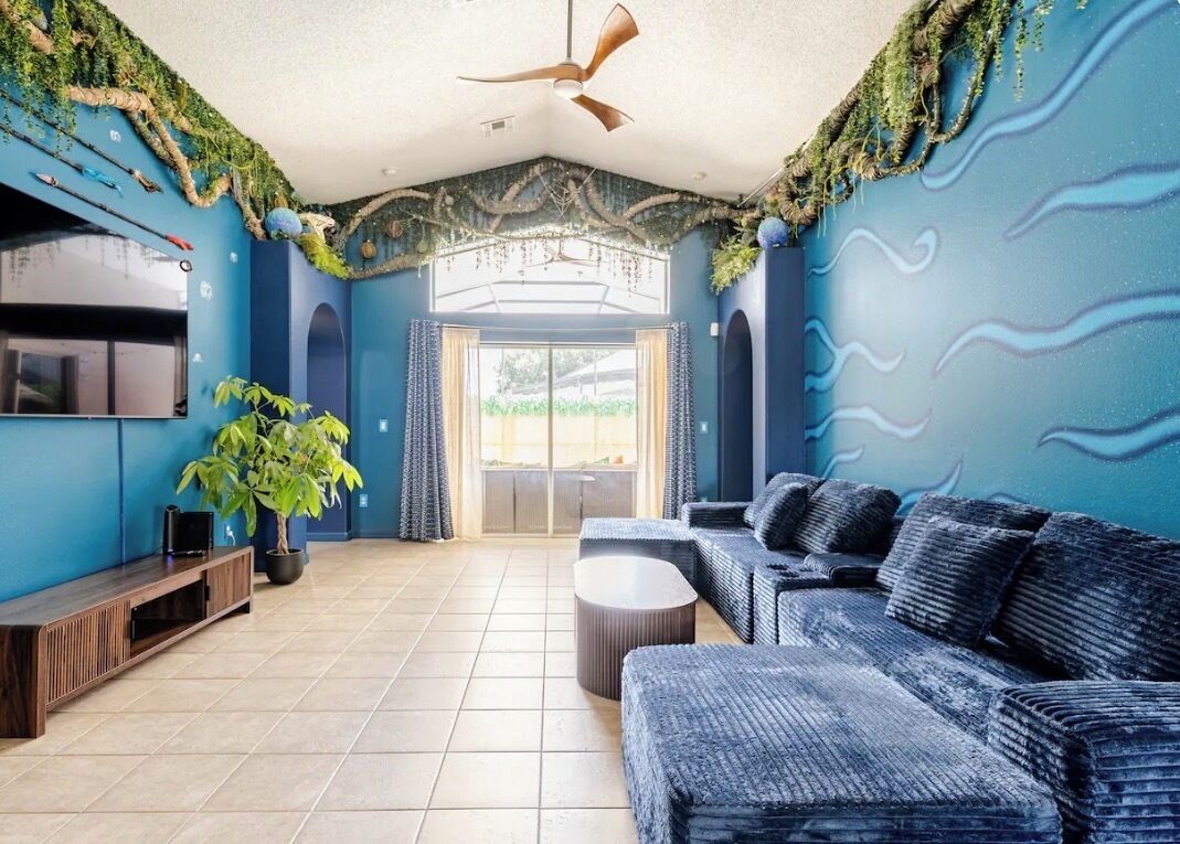 A room with a blue theme, a ceiling fan, a sofa, a TV, and a plant.