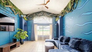 Quarto com decoração azul, ventoinha no teto, sofá, televisão e planta.