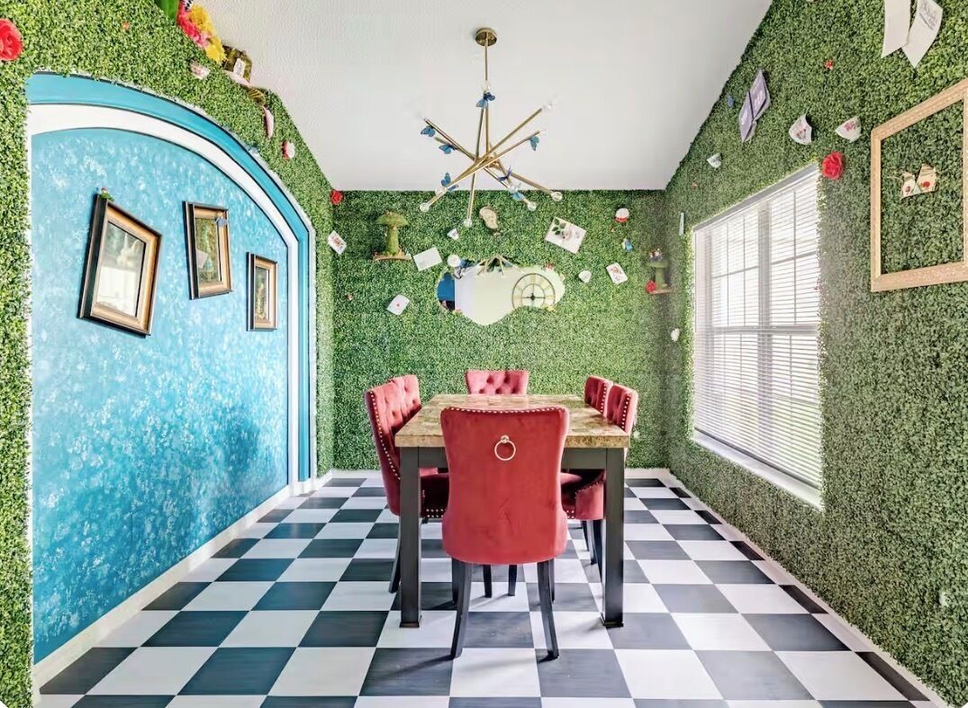 Un comedor con piso a cuadros, una mesa con sillas tapizadas en rojo y una pared decorada con plantas y cuadros.
