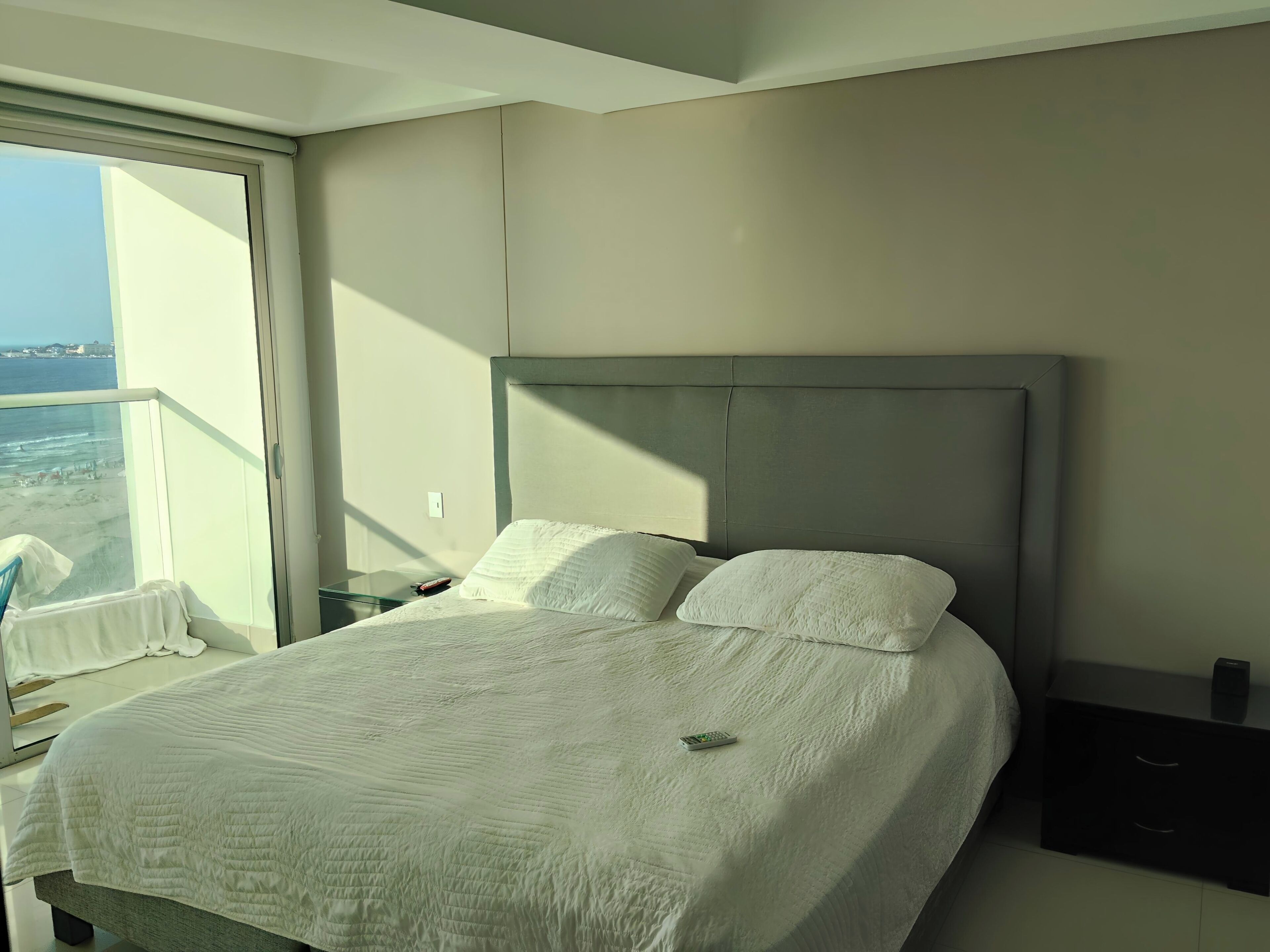 A hotel room with a large bed, a nightstand, a glass table, and a view of the sea.