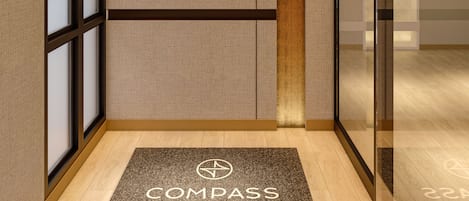 Um hall de hotel com design moderno, com balcão de recepção, divisória de vidro e tapete de entrada com o nome do hotel.