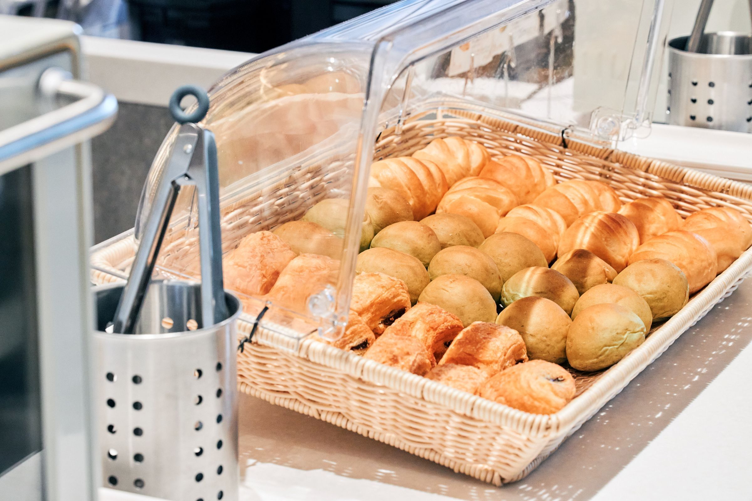 A buffet with a wicker basket containing various baked goods, including croissants and rolls, under a clear plastic cover.