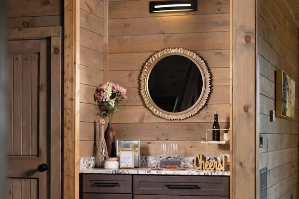 A wooden-paneled bathroom with a round mirror, a vase with flowers, and a shelf with a bottle and glasses.