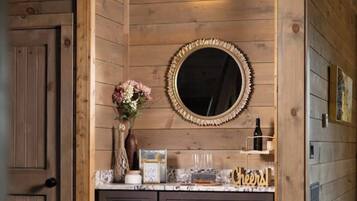 A wooden-paneled bathroom with a round mirror, a vase with flowers, and a shelf with a bottle and glasses.