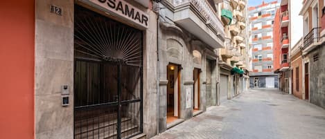 Una calle estrecha con un edificio identificado como 'ROSAMAR' a la izquierda, que presenta una gran puerta metálica y una entrada pequeña.