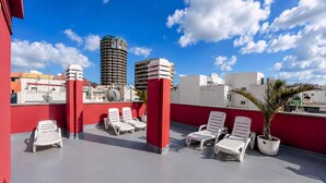 A rooftop terrace with white lounge chairs, red pillars, and a view of surrounding buildings. - GuestReady - Rosamar P Apartments (Las Palmas de Gran Canaria)