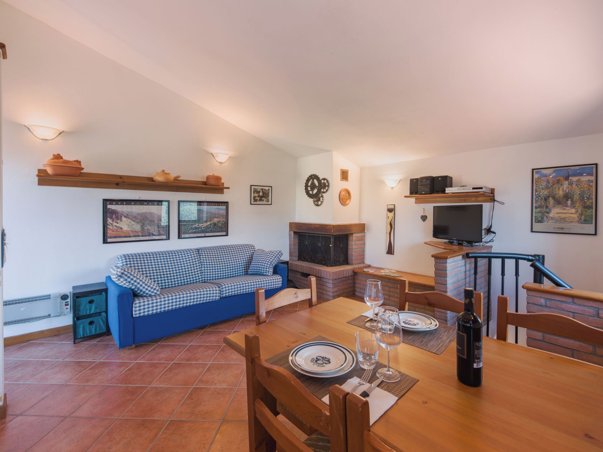 Un salón acogedor con un sofá azul a cuadros, una chimenea, una mesa de comedor lista para dos personas y un televisor montado en la pared.