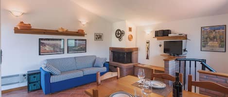 Un salón acogedor con un sofá azul a cuadros, una chimenea, una mesa de comedor lista para dos personas y un televisor montado en la pared.