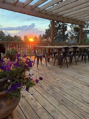 Una terraza de madera con una mesa y sillas, rodeada por una barandilla de madera, y una puesta de sol al fondo.