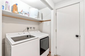 A laundry area with a white washing machine, a shelf with various items, and a closed door.