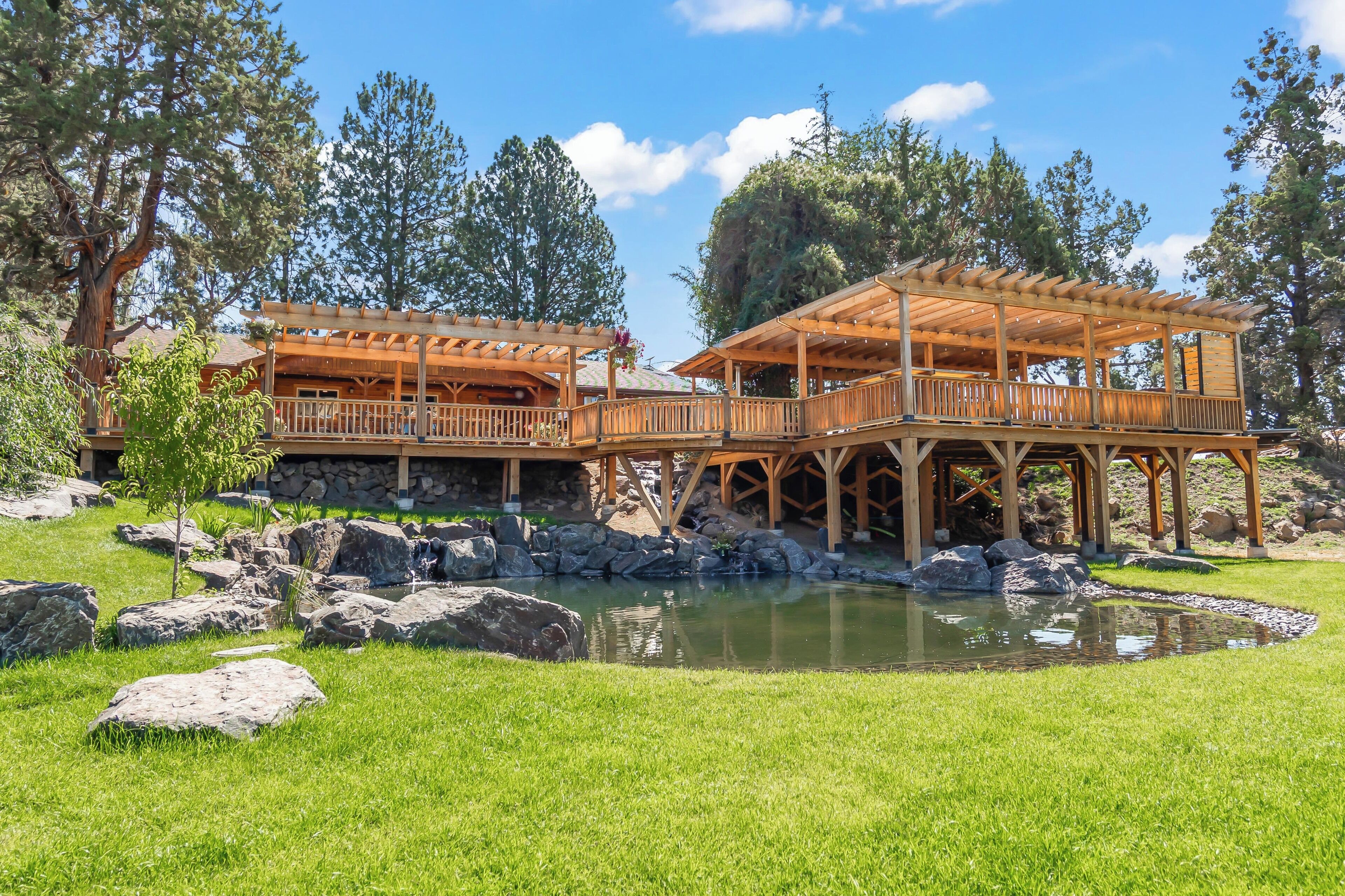 A wooden gazebo with a stone foundation, surrounded by a pond and large rocks.