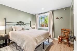 Un dormitorio con cama, mesita de noche, lámpara, ventana con cortinas y una silla.