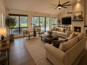 A spacious living room with a fireplace, large windows, and a ceiling fan. - Luxury Oak Escape l Hot
Tub & Nature &Game Room (Bulverde)