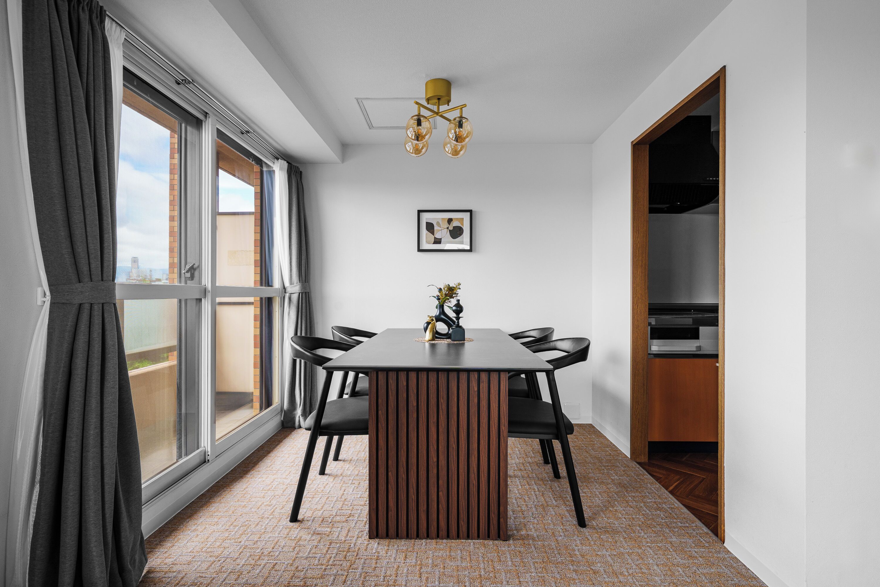 A modern dining area with a wooden table and chairs, large windows with curtains, a ceiling light fixture, and a framed picture on the wall.