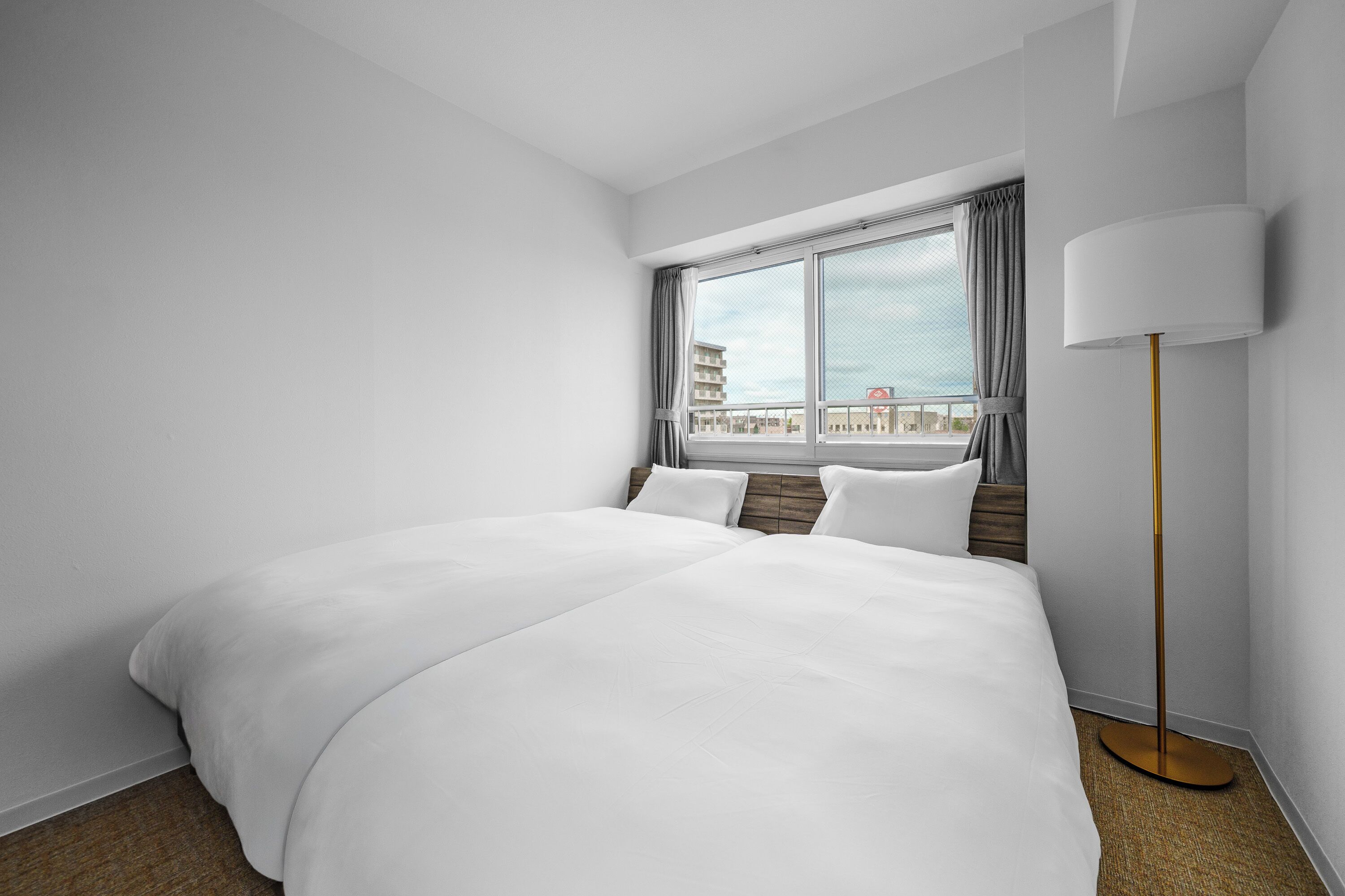 A hotel room with a large bed, a standing lamp, and a window with a view of buildings.