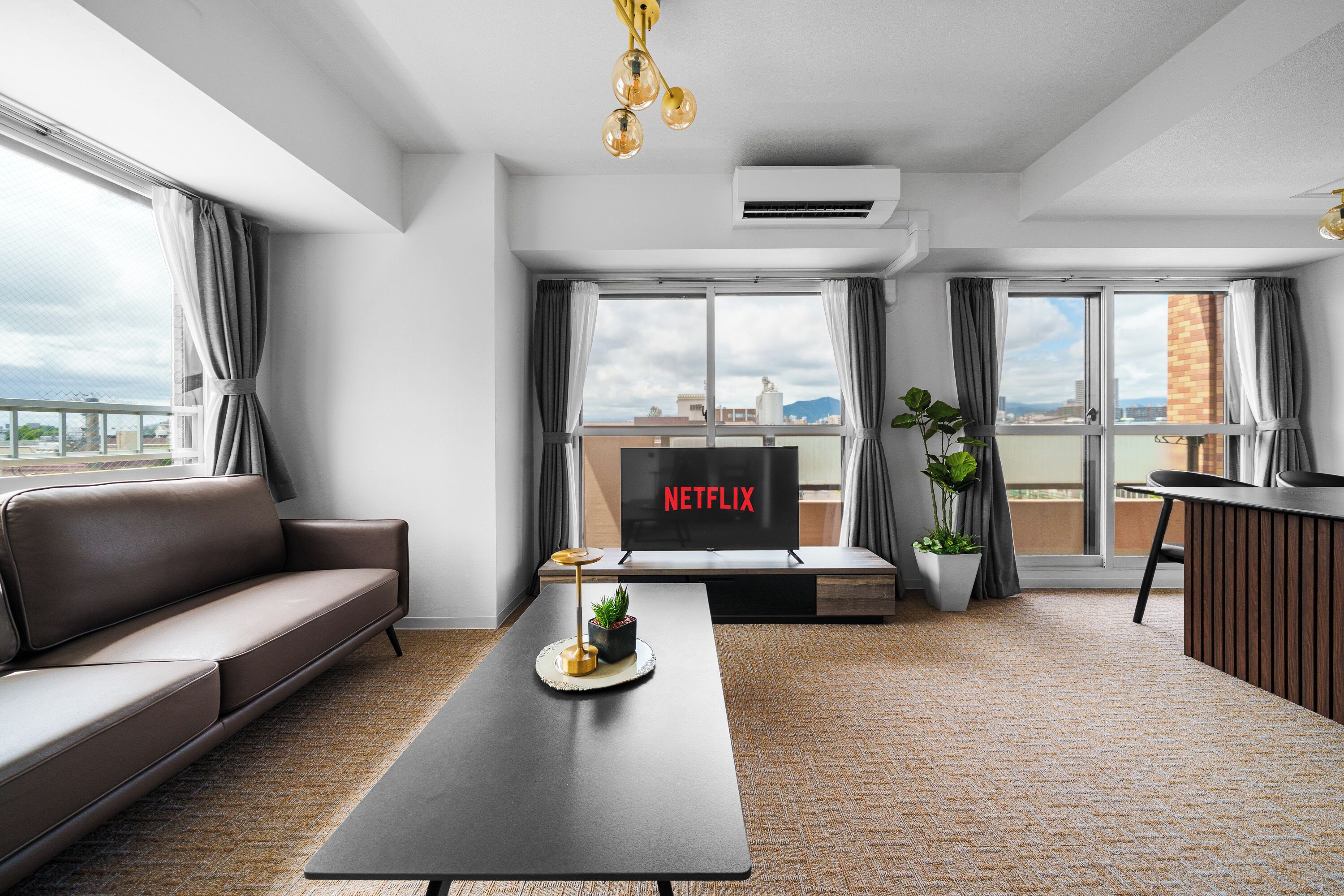 A modern living room with a sofa, a coffee table, a TV displaying 'NETFLIX', and a balcony with a view.