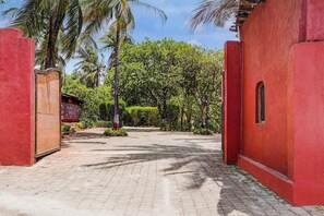 A paved pathway leading to a red building with a small window, surrounded by lush greenery and palm trees.