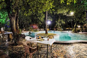 An outdoor pool area with a table and chairs, surrounded by trees and a small building.