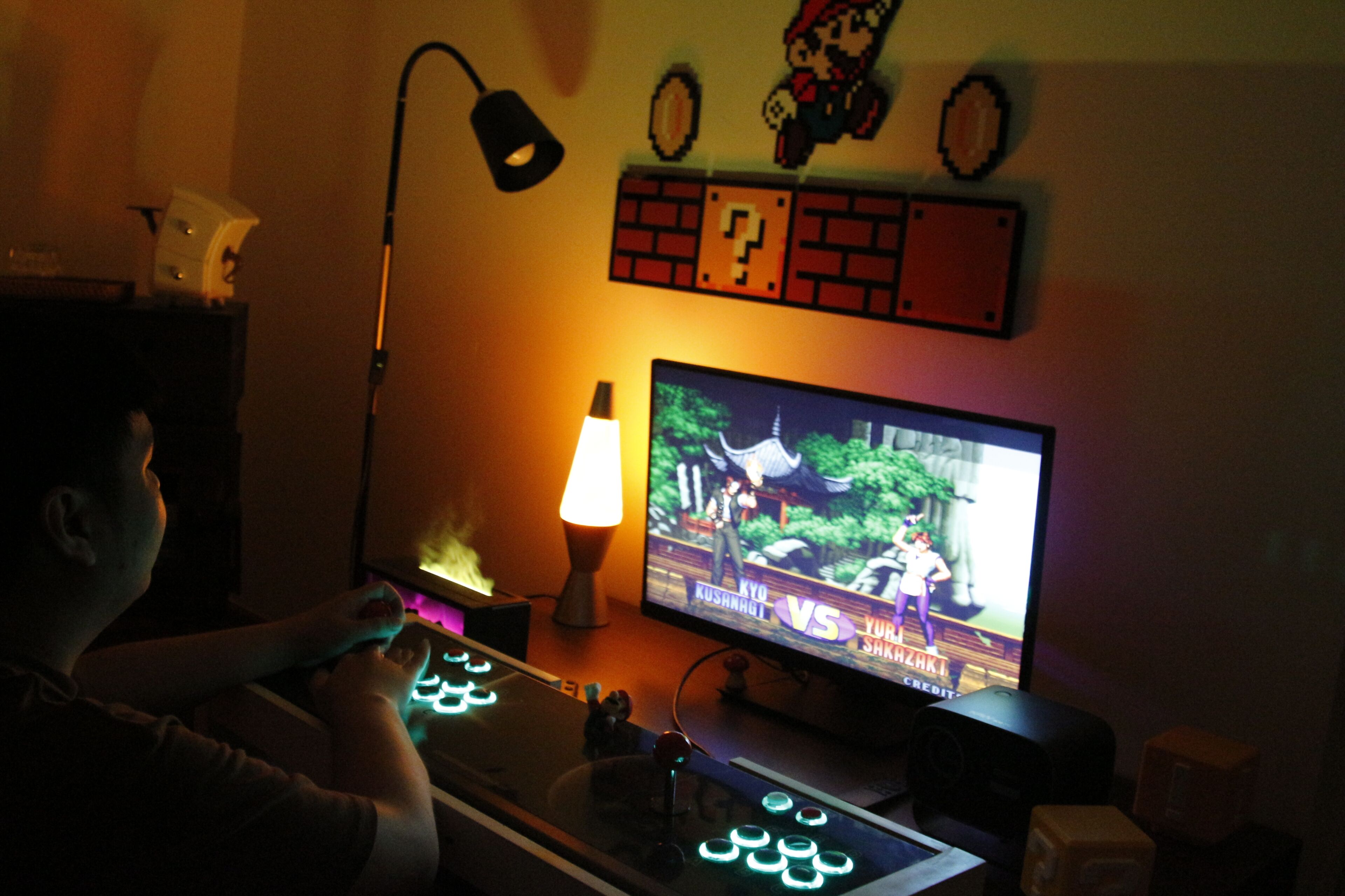 A person playing a video game with a TV mounted on a wall, a lamp, and a keyboard with illuminated keys.