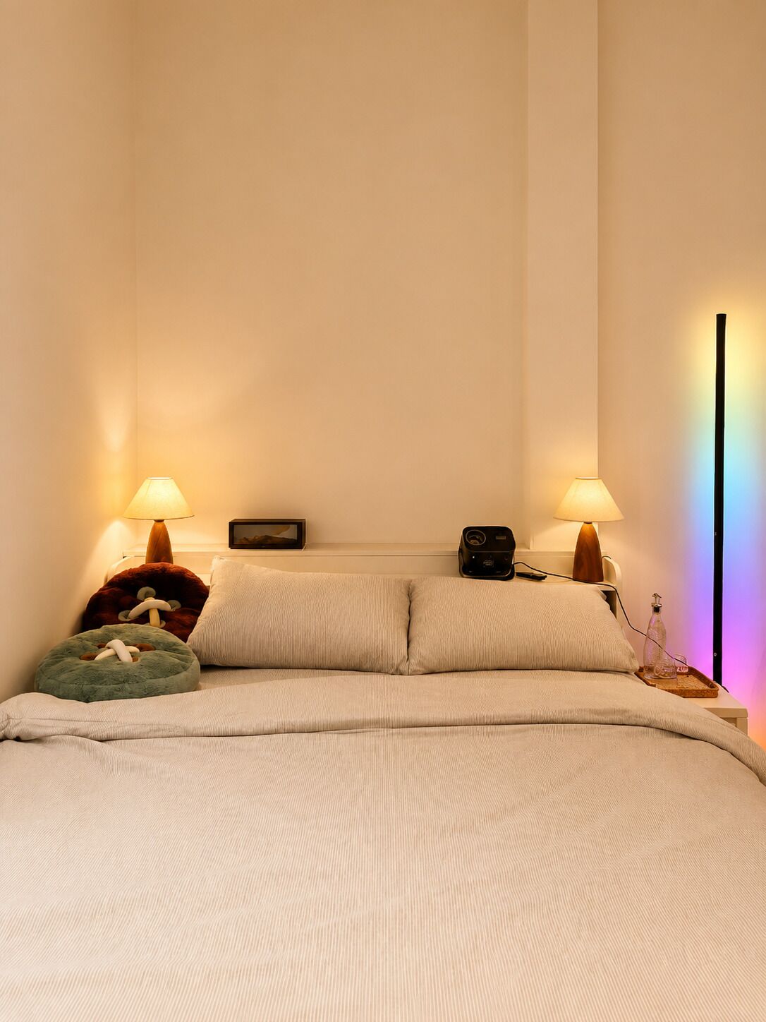 A neatly made bed with two pillows, two bedside lamps, a small table with a radio, and a wall-mounted shelf with a picture frame.