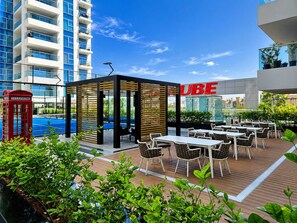 Un espace extérieur aménagé avec des tables et des chaises, un téléphone public rouge et un bâtiment moderne avec une enseigne « UBE » en arrière-plan.