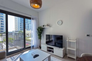 Ein modernes Wohnzimmer mit großem Fernseher, einem weißen Couchtisch, einer Pflanze und einem Balkon mit Blick auf Gebäude.