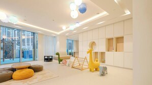 A modern living room with a large window, a sofa, a play area with a giraffe and elephant toy, and a bookshelf. - Luxury 1BR at Grande | Cityscape Views, Downtown (Dubai)