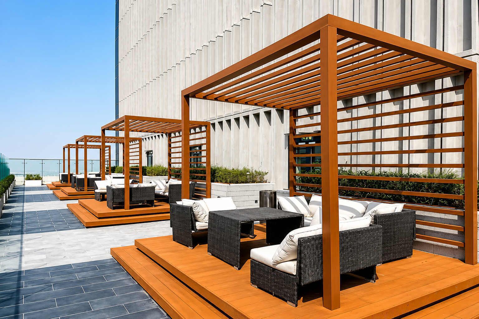 Zonas de estar al aire libre con muebles de mimbre y pérgolas de madera, frente a la fachada de un edificio moderno.
