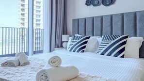Ein Hotelzimmer mit Bett, Kissen, Nachttisch, Lampe und Blick auf ein Gebäude durch ein Fenster.