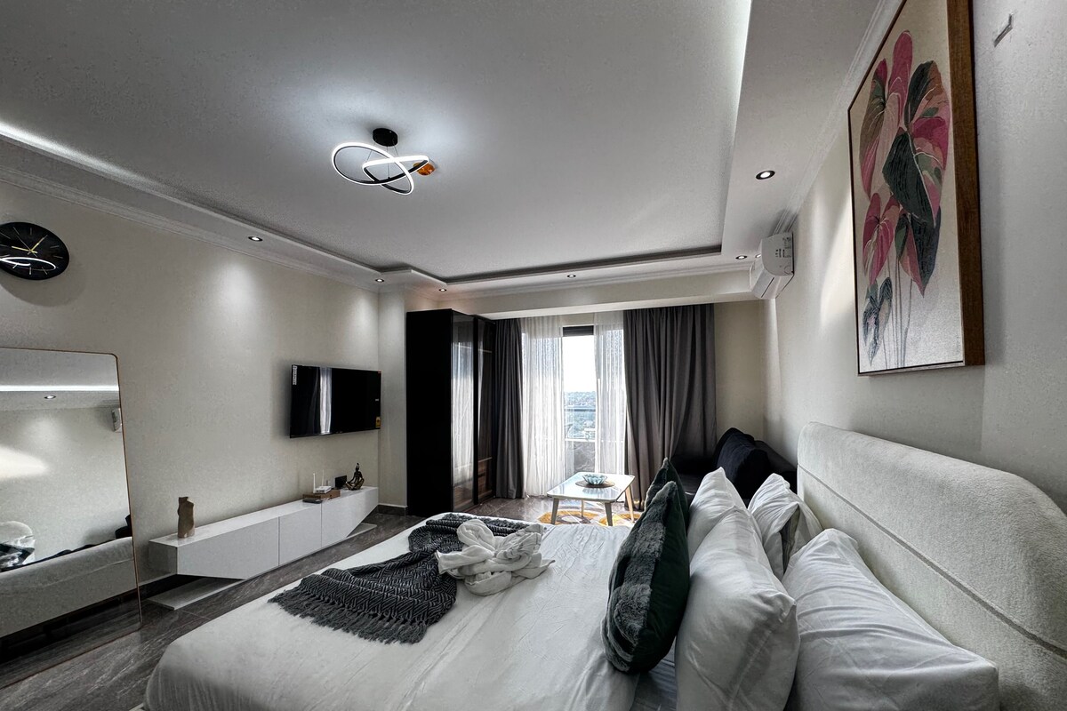 A modern hotel room with a large bed, a flat-screen TV, a bedside table, and a view of the city through the window.