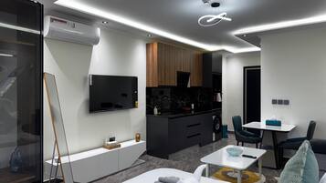 A modern living room with a flat-screen TV, a dining area, and a kitchen with dark cabinetry.