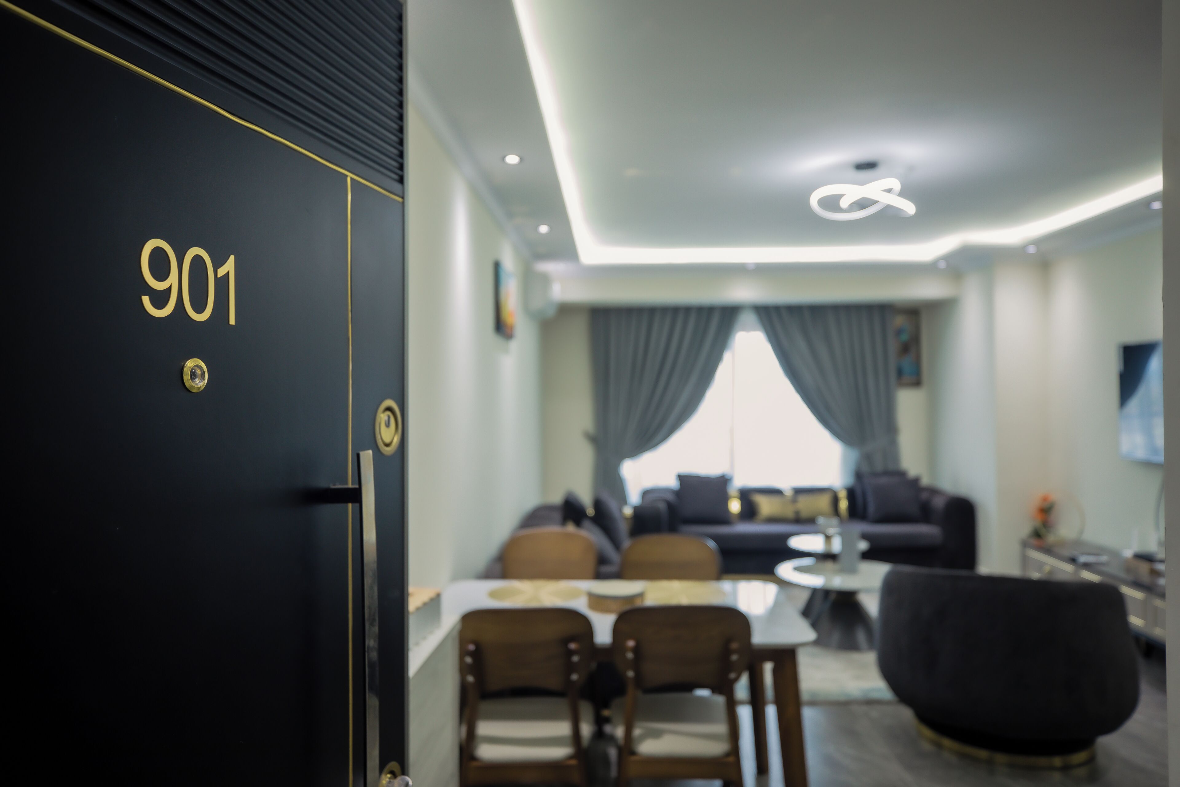 A modern interior with a black door marked '901', a dining area with chairs and a table, a sofa, and a large black armchair.