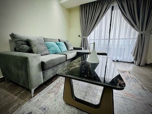 A modern living room with a grey sofa, a glass coffee table, and a view through large windows with curtains.
