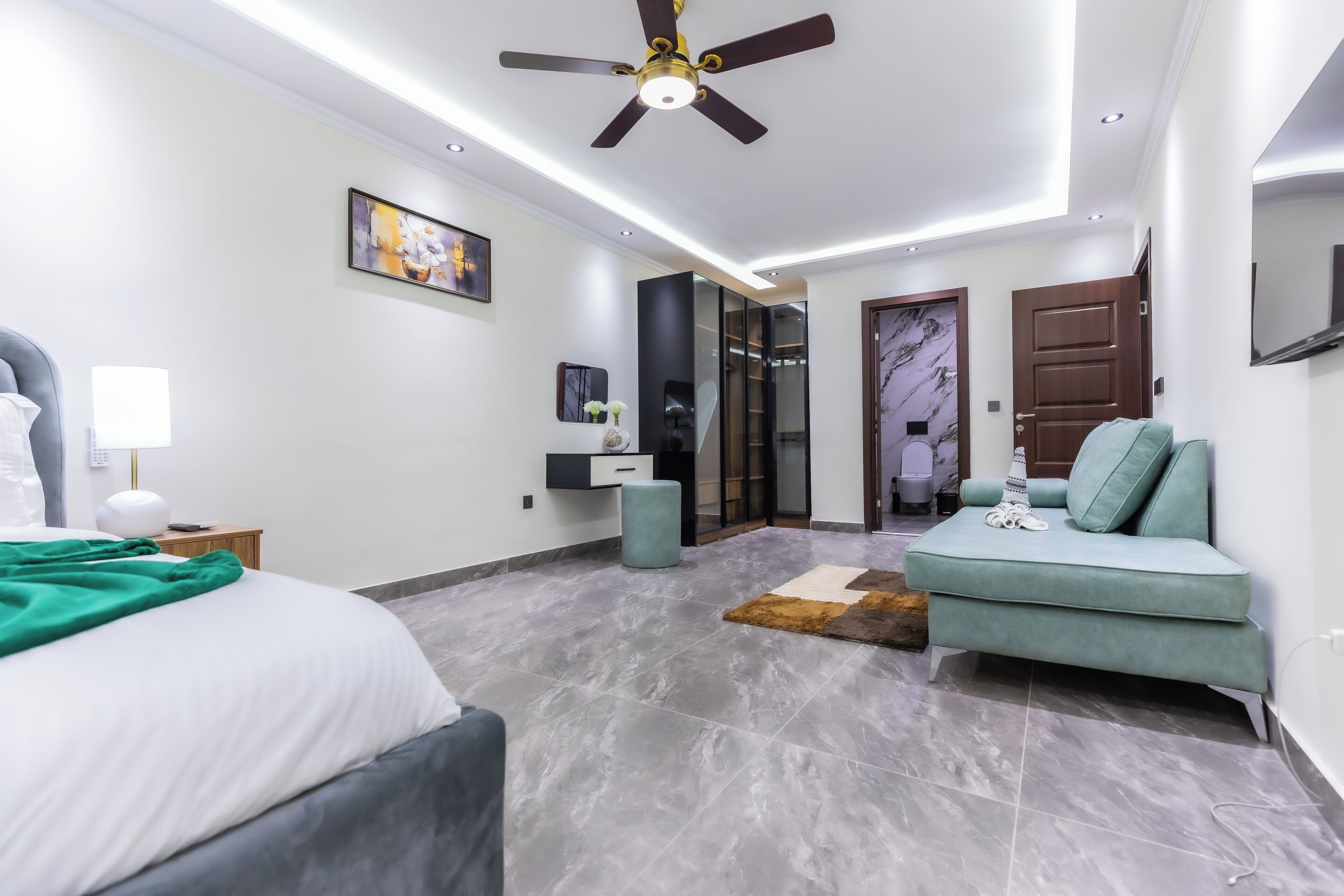 A modern bedroom with a bed, a seating area, a ceiling fan, and a door leading to a bathroom.