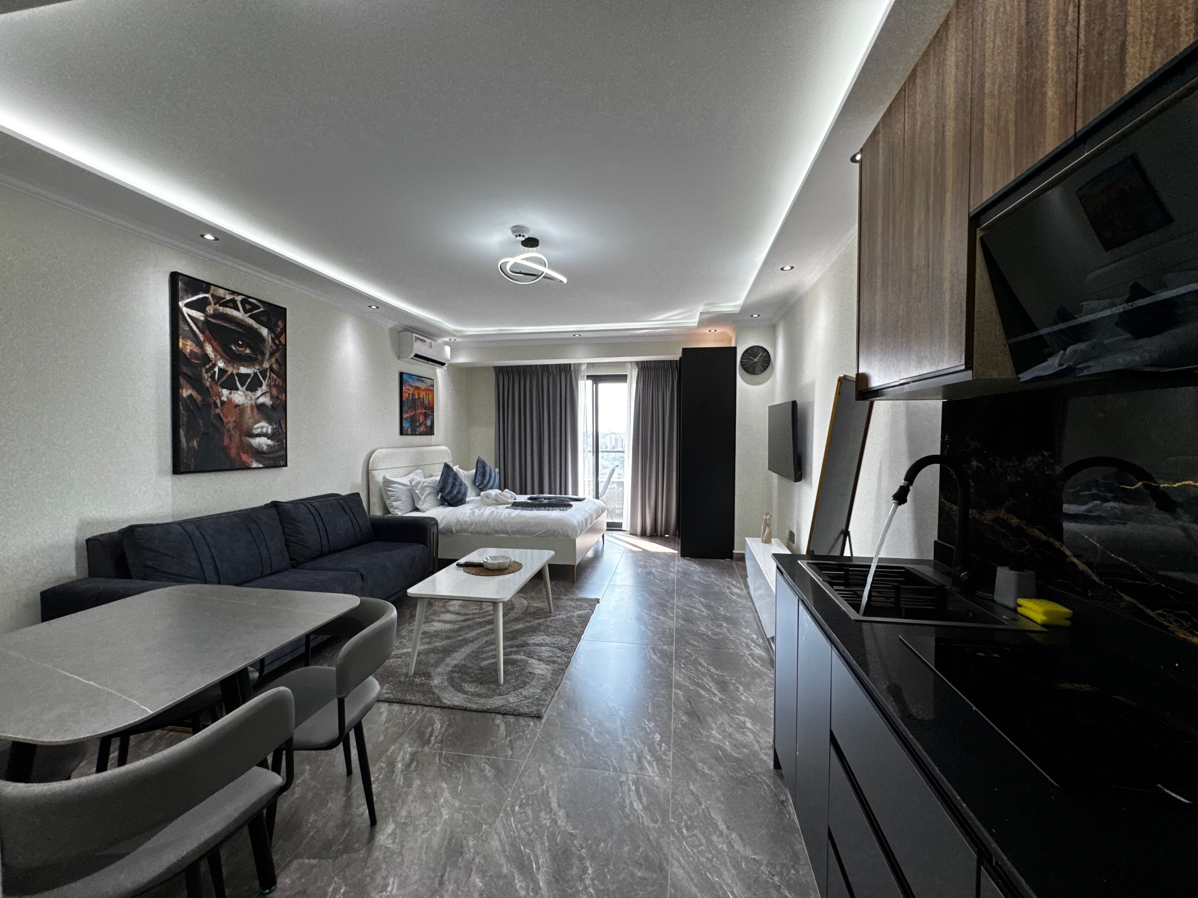 A modern hotel room with a dining area, a sofa, a bed, a kitchen area, and artwork on the walls.
