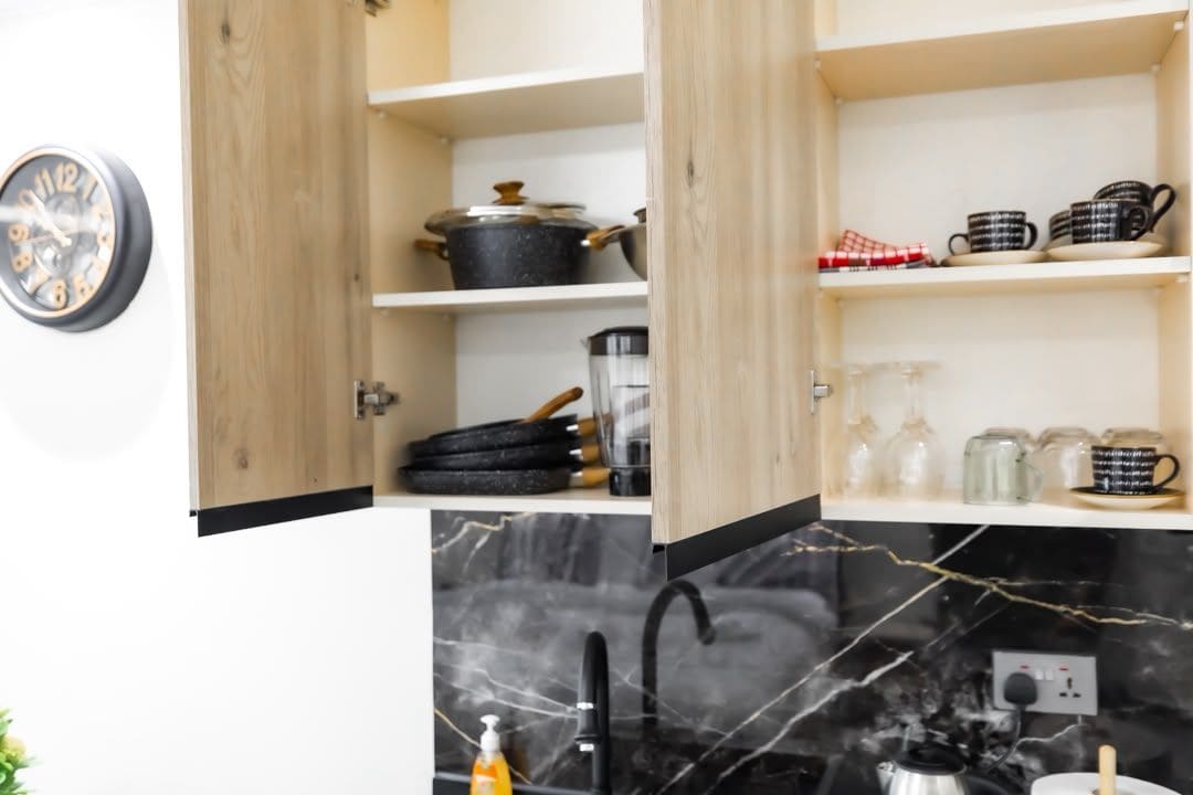 Een keuken met open houten kastjes, een zwart marmeren achterwand en diverse keukengerei.