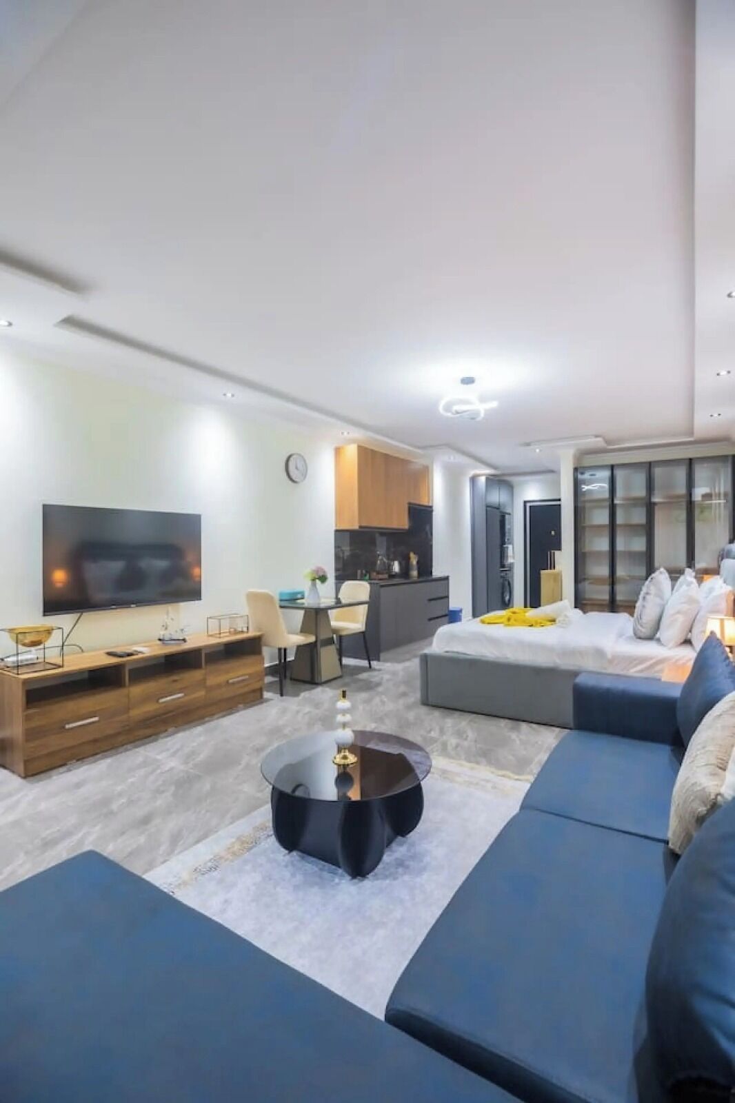 A modern living room with a flat-screen TV, a coffee table, a sofa, a desk with a chair, and a bed with pillows.