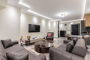 A modern living room with a flat-screen TV, a sofa, a coffee table, and a dining area with chairs. - Floria 807B (Accra)