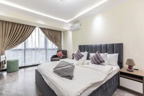 A modern hotel room with a large bed, a chair, a nightstand, and a view of the city. - Floria 807B (Accra)