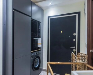 A compact laundry room with a washing machine and dryer, a door with a lock, and a small chair. - Floria 807B (Accra)